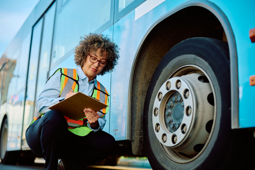 Woman in safety vest inspecting tire on a commercial bus, writing on a clipboard