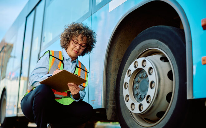 Woman in safety vest inspecting tire on a commercial bus, writing on a clipboard