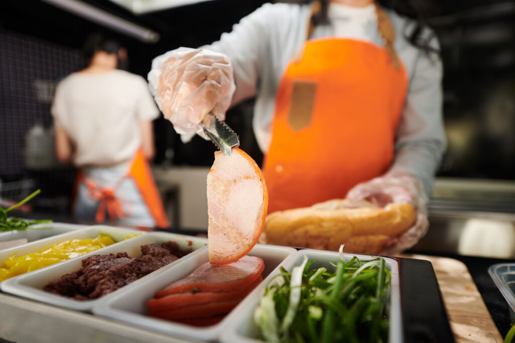 Person in commercial kitchen, wearing gloves, holding sliced meat with tongs