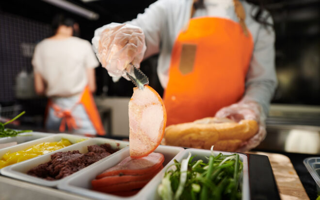 Person in commercial kitchen, wearing gloves, holding sliced meat with tongs