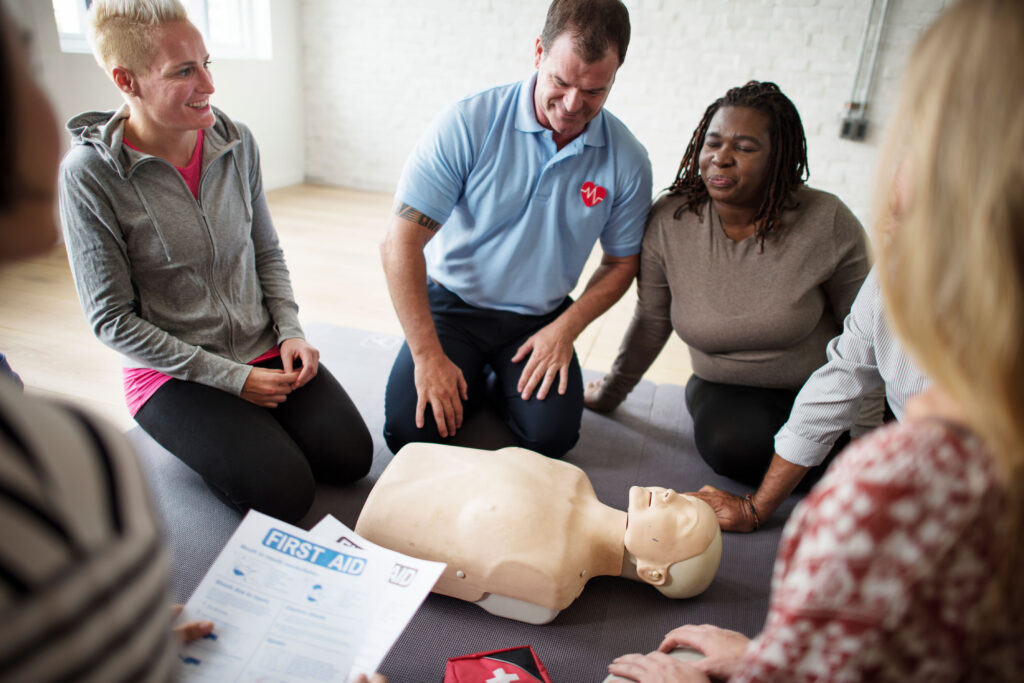 Group of people around a CPR training dummy, one person in foreground is holding papers labeled "first aid"