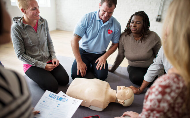 Group of people around a CPR training dummy, one person in foreground is holding papers labeled "first aid"