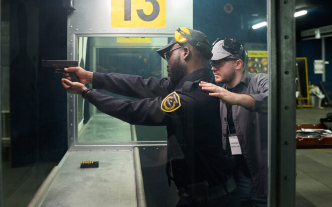 A firearms instructor helps a security guard line up a shot with a handgun in an indoor firearms training range.