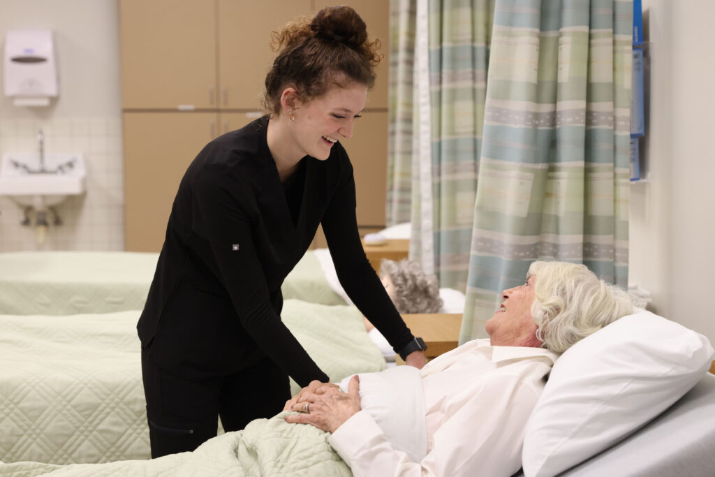 Nursing Assistant student smiling at an elderly woman in a medical bed