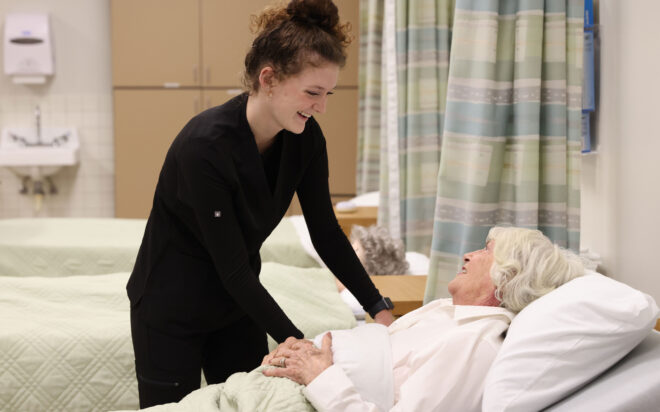Nursing Assistant student smiling at an elderly woman in a medical bed